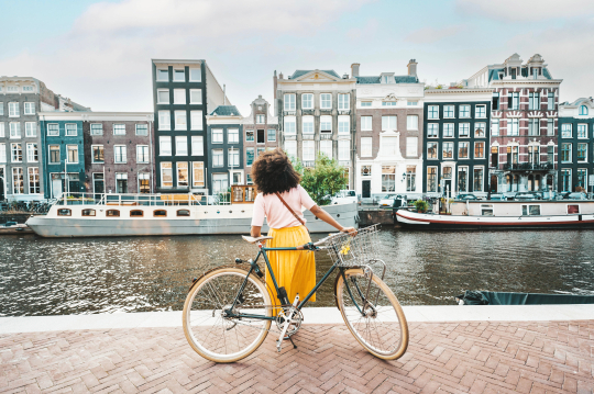 Frau mit gelben Rock und weißem Shirt lehnt an ihrem Fahrrad mit Blick auf eine Häuserreihe an einer Gracht in Amsterdam